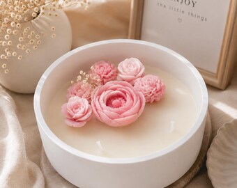 May include: A round white candle container filled with cream-colored wax, featuring a cluster of pink floral-shaped wax decorations and two wicks. The arrangement includes several small rose-like blossoms and a larger central flower.