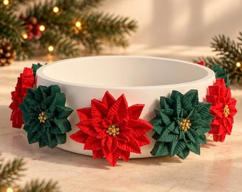 May include: A round white bowl decorated with alternating red and dark green poinsettia flowers around the exterior. Each flower features a cluster of small gold beads at its center, creating a festive appearance on the smooth white surface.