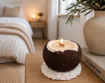 May include: A dark brown, textured coconut-shaped candle with a lit wooden wick, resting on a white circular base featuring a raised floral pattern. The candle is positioned next to a beige vase and a book with the text EAT DRINK NAP printed on it.