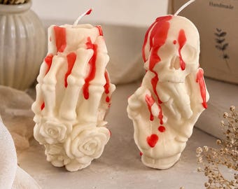 May include: Two off-white wax candles shaped like skeletal hands and a skull, featuring red wax drips resembling blood. One candle depicts a hand with sculpted roses at the base, while the other shows a skull resting on a hand, both with wicks.