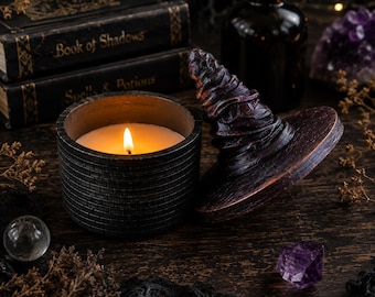 May include: A dark, cylindrical candle container with a brick-patterned exterior and a lit candle inside. Beside it lies a matching dark, conical lid shaped like a pointed hat with a textured, swirling surface and a wide, circular brim.