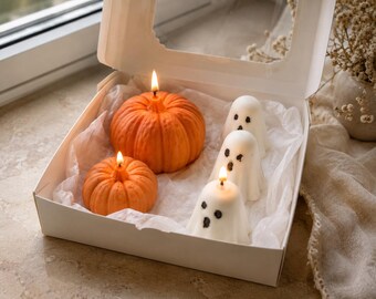 May include: A white cardboard box with a clear window contains two orange pumpkin-shaped candles and three white ghost-shaped candles. Each candle is lit with a small flame, resting on white tissue paper inside the open container.