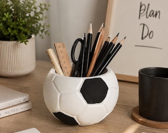 May include: A white and black soccer ball shaped desk organizer holding several black pencils, a pair of black scissors, a wooden ruler, and a light-colored pen. The container features the classic hexagonal and pentagonal pattern of a soccer ball.
