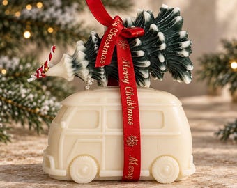 May include: A cream-colored van-shaped candle topped with a dark green and white Christmas tree candle. A red ribbon with the gold text Merry Christmas is tied around the van and tree. A small red and white striped string is attached to the tree.