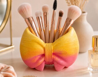 May include: A bow-shaped makeup brush holder with a yellow-to-pink gradient finish, containing five makeup brushes with rose gold ferrules and light-colored bristles. The holder has a textured surface and is shaped like a tied ribbon.