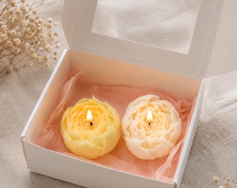 May include: Two lit flower-shaped candles, one yellow and one cream, nestled inside a white cardboard gift box with a clear window lid. The candles rest on light pink tissue paper, and each features a small, flickering flame at its center.