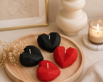 May include: Four geometric heart-shaped candles arranged on a round wooden tray. Two candles are solid black and two are solid red. Each candle features a faceted surface design and a single white wick centered at the top of the heart shape.
