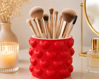 May include: A cylindrical makeup brush holder in bright red featuring a repeating embossed heart pattern on the exterior. The holder is filled with a collection of various makeup brushes with light brown bristles and metallic gold-toned handles.