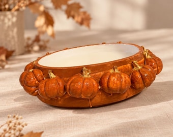 May include: An oval-shaped ceramic bowl with a smooth white interior and a textured exterior featuring a series of sculpted, glossy orange pumpkins arranged in a continuous row around the entire outer rim of the vessel.