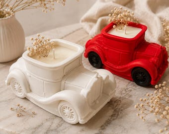 May include: Two car-shaped candles, one matte white and one bright red, each featuring a central wick and a small sprig of dried baby's breath flowers placed inside the open roof area. The candles have black wheels and detailed car body molding.