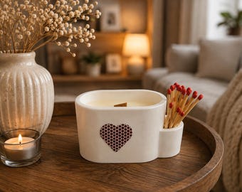 May include: A white oval ceramic candle vessel with a wooden wick and a built-in side compartment holding a cluster of wooden matches with red tips. The front of the vessel features a heart-shaped strike pad with a dark red honeycomb pattern.