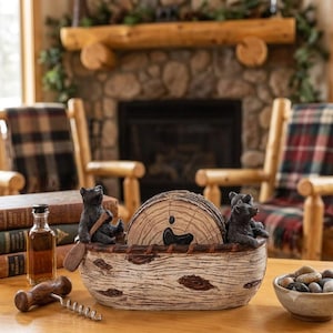 May include: A decorative canoe-shaped holder with a textured birch bark finish, featuring two black bear figurines at the ends and a set of circular wood-grain coasters stored inside. The holder is crafted with realistic bark details and brown accents.