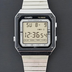 Puede incluir: Un reloj digital Casio TC-500 de tono plateado con correa de eslabones metálicos. La esfera rectangular presenta un bisel negro y una pantalla digital clara que muestra la hora 12:36:54, la fecha SU 8-9 y símbolos de calculadora abajo.