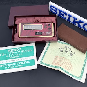 May include: A gold-tone Seiko Quartz digital pocket alarm clock with a maroon patterned face, housed in a maroon box. Includes a white instruction manual with green text, a warranty card, a brown protective sleeve, and a white Seiko display sign.