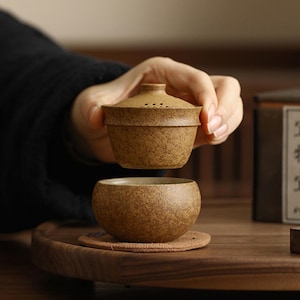 Può includere: Un set da tè in ceramica marrone testurizzata composto da una piccola ciotola arrotondata e un coperchio abbinato con una serie di piccoli fori. Il coperchio è tenuto sopra la ciotola, rivelando la sua finitura maculata e terrosa.
