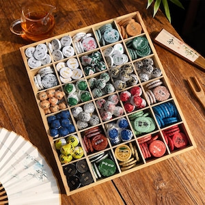 Peut inclure: Une boîte de rangement carrée en bois avec de multiples compartiments remplis de petits gâteaux de thé ronds et emballés individuellement, de couleurs variées dont blanc, vert, bleu, rouge, jaune et noir, avec des étiquettes et du texte imprimés.