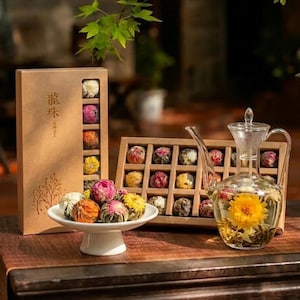 Puede incluir: Una tetera de cristal transparente que contiene una flor de té amarilla en flor, acompañada de un cuenco blanco con pedestal lleno de bolas de té de colores. Una caja marrón alta con caracteres chinos y una caja de rejilla con té están cerca.