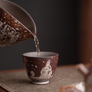 May include: A dark brown ceramic tea cup with a speckled texture and white etched figures of seated deities. A stream of clear liquid is being poured from a matching ceramic pitcher into the cup, which has a light-colored interior rim.