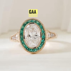 Peut inclure: Une bague en or avec une grande pierre centrale ovale entourée d'un halo de petites pierres vertes taille baguette. L'anneau est orné de petites pierres claires et présente un bord milgrain. Le texte GAA GEMARTBYALI est visible au-dessus.