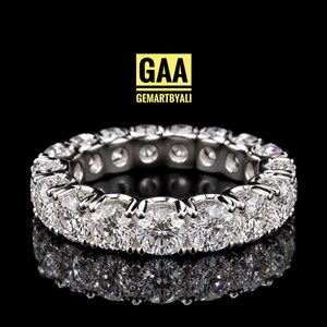 Peut inclure: Une bague alliance en métal argenté ornée d'une rangée continue de pierres rondes serties dans des griffes. Au-dessus de la bague, un logo carré jaune affiche le texte noir GAA et le texte plus petit GEMARTBYALI.
