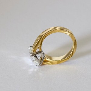 Peut inclure: Une bague en or dot&eacute;e d'un anneau textur&eacute; &agrave; rainures et d'une pierre pr&eacute;cieuse transparente &agrave; facettes mont&eacute;e sur un sertissage &agrave; griffes argent&eacute;. L'anneau pr&eacute;sente un motif de lignes verticales distinct qui fait le tour complet, contrastant avec l'int&eacute;rieur lisse et poli.