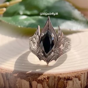 Peut inclure: Une bague argentée avec une pierre noire centrale taillée en marquise fixée par quatre griffes. L'anneau présente un travail de métal complexe avec des motifs de feuilles et des bords pointus. Le texte GAAGemArtByAli est visible au-dessus.