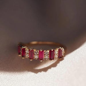 May include: A gold ring featuring a row of alternating rectangular baguette-cut red rubies and small round white diamonds. The band is crafted from a polished gold metal, showcasing a series of five vibrant red gemstones set between pairs of clear, sparkling white stones in a continuous horizontal arrangement.