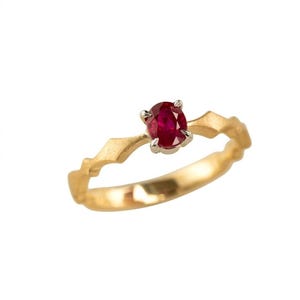 Puede incluir: Un anillo de oro con un rubí rojo de talla oval engastado en una montura de plata de cuatro garras. La banda presenta un diseño geométrico, angular y texturizado que se estrecha hacia el centro, contrastando con el acabado dorado pulido.
