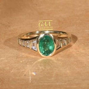 Peut inclure: Une bague en or avec une émeraude verte centrale de taille ovale dans un serti clos, flanquée de trois pierres claires de taille baguette de chaque côté de l'anneau. Le texte GAA GemArtByAli est visible au-dessus de la bague en jaune.