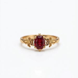 May include: A gold ring featuring a central oval-cut deep red gemstone held by four prongs. The band displays intricate metalwork on both sides of the stone, characterised by symmetrical, leaf-like scroll patterns that extend along the ring shank.