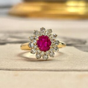 May include: A gold ring featuring a round, vibrant red gemstone at the centre, surrounded by a halo of twelve small, clear, round-cut diamonds arranged in a floral pattern. The band is a smooth, polished gold metal that tapers towards the setting.
