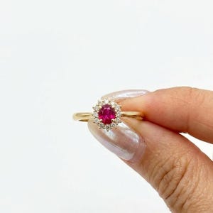 Könnte beinhalten: Ein Goldring mit einem zentralen, oval geschliffenen, leuchtend roten Edelstein, der von einem Kranz aus kleinen, klaren, rund geschliffenen weißen Steinen umgeben ist. Die Ringschiene besteht aus poliertem, glattem Goldmetall.