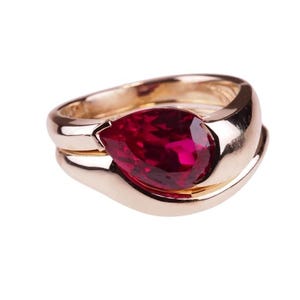 Puede incluir: Un anillo de oro rosa pulido con una piedra preciosa de color rojo intenso en forma de pera engastada horizontalmente. La banda tiene un diseño liso, ancho y curvo que envuelve la base de la piedra, creando una silueta metálica fluida.
