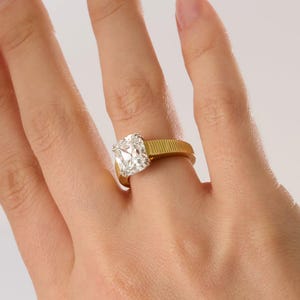 Peut inclure: Une bague en or avec un anneau textur&eacute; et nervur&eacute;, orn&eacute;e d'une grande pierre transparente taille coussin mont&eacute;e sur un sertissage argent&eacute;. La bague est port&eacute;e au doigt, mettant en valeur le contraste entre l'anneau jaune et la pierre.