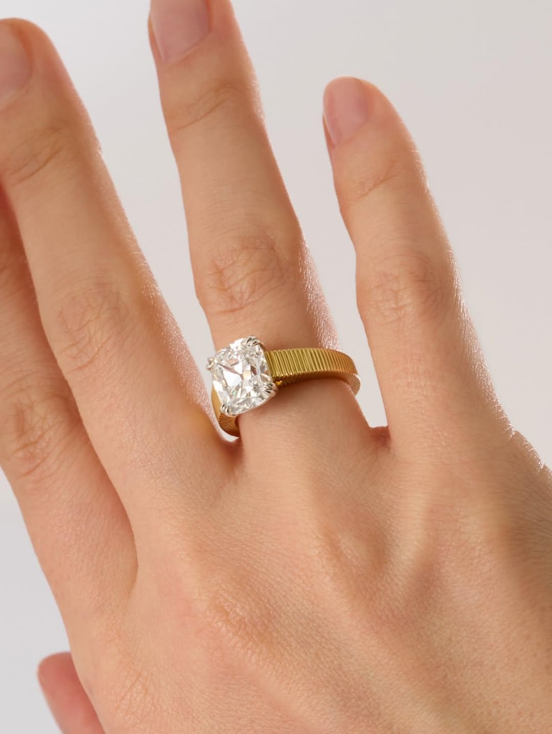 Peut inclure: Une bague en or avec un anneau textur&eacute; et nervur&eacute;, orn&eacute;e d'une grande pierre transparente taille coussin mont&eacute;e sur un sertissage argent&eacute;. La bague est port&eacute;e au doigt, mettant en valeur le contraste entre l'anneau jaune et la pierre.