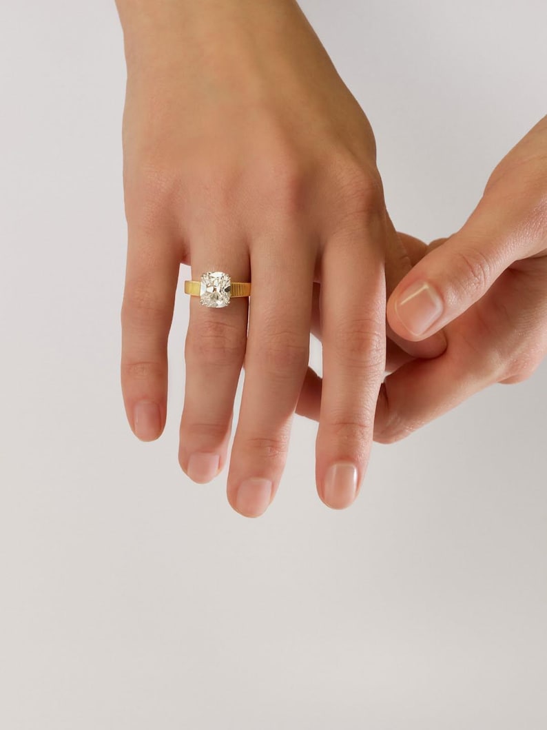 Peut inclure: Une bague en or orn&eacute;e d'une grande pierre transparente de taille coussin rectangulaire au centre. L'anneau de la bague pr&eacute;sente une finition distincte, verticalement nervur&eacute;e ou textur&eacute;e, contrastant avec la surface lisse et facett&eacute;e.