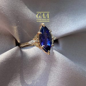 Peut inclure: Une bague en or avec une grande pierre précieuse bleu foncé taille marquise au centre, flanquée de plus petites pierres claires taille marquise et de détails métalliques en forme de feuilles. Le texte GAA GemArtByAli est visible au-dessus.