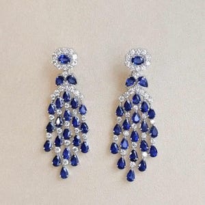 Puede incluir: Un par de pendientes tipo candelabro con zafiros azules de corte ovalado en la parte superior, rodeados de pequeñas piedras claras. Debajo, múltiples hilos de zafiros azules de corte de pera y piedras claras cuelgan en un diseño en cascada.