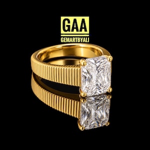 May include: A gold ring featuring a rectangular radiant-cut clear gemstone held by four prongs. The band has a distinct vertical ribbed texture. Above the ring, the text GAA GEMARTBYALI is displayed in black letters on a yellow square background.