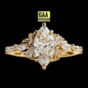 Peut inclure: Une bague en or avec une grande pierre centrale taille marquise, entourée de pierres plus petites taille poire et ronde sur un anneau décoratif. Le texte GAA GEMARTBYALI est visible au-dessus de la bague sur fond noir.
