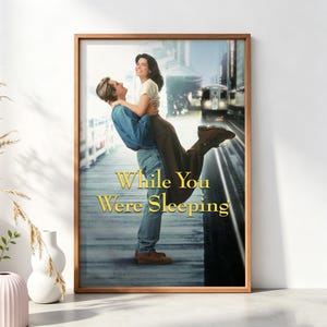 以下が含まれることがあります： 映画『あなたが寝てる間に』の額入りポスター。青いシャツとジーンズを着た人物が、白いトップスと茶色のスカートを着た人物を駅のホームで持ち上げている様子が描かれており、背景には列車、中央には金色の文字がある。