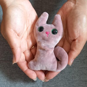 May include: A small, soft, mauve-coloured plush cat toy with a smooth, velvety texture. The cat features two pointed ears, a curled tail, and two large, round, dark green glittery eyes. A small, simple pink stitched nose is centred on the face.