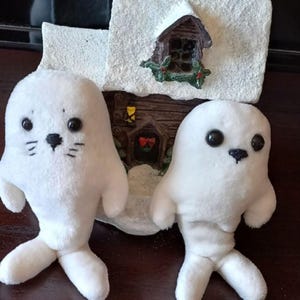 May include: Two white plush seal toys with black plastic eyes and stitched facial features. The left seal has a stitched nose and whiskers, while the right seal has a simple stitched nose. Behind them sits a small, snow-covered cabin figurine.