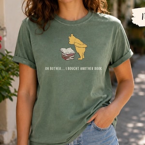 May include: A moss green short-sleeve t-shirt featuring a graphic of a yellow bear standing next to a stack of books. Below the graphic, the text reads: OH BOTHER... I BOUGHT ANOTHER BOOK. The shirt has a crew neckline and a relaxed fit.