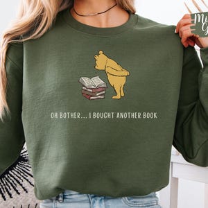 May include: A military green crewneck sweatshirt featuring a graphic of a yellow bear looking at a stack of books. Below the graphic, the text reads: OH BOTHER... I BOUGHT ANOTHER BOOK. The sweatshirt has long sleeves and a ribbed collar, cuffs, and hem.