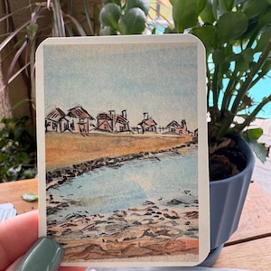 May include: A rectangular card featuring a watercolor illustration of a coastal landscape. The scene depicts a sandy beach in the foreground, a calm blue body of water, and a row of small houses with brown roofs under a pale blue sky.