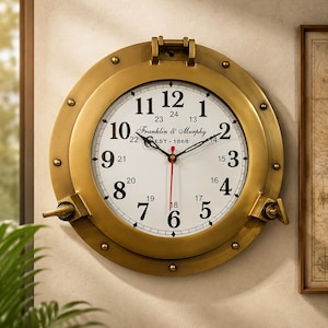 May include: A round wall clock with a brass-colored porthole frame featuring rivets and two side latches. The white face displays black Arabic numerals from 1 to 12, smaller 13 to 24 hour markers, and the text Franklin & Murphy EST 1868.