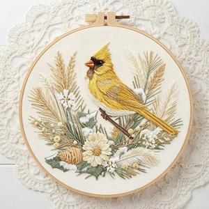May include: A circular embroidery hoop containing a fabric panel with a detailed thread-stitched yellow cardinal bird perched on a branch, surrounded by embroidered wheat stalks, white flowers, pine needles, and a small pinecone.