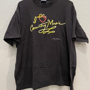 May include: Black t-shirt with a yellow and red graphic that says "I Love Country Music" with a crown on the heart. The graphic is in a handwritten style. The text "Brenda Lee (In My Own Words)" is printed below the graphic.