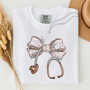 Peut inclure: Un t-shirt blanc à manches courtes avec un imprimé graphique représentant un stéthoscope en forme de nœud. Le nœud présente un motif floral bleu clair et rose avec des brins de lavande. L'étiquette Comfort Colors est visible au col.